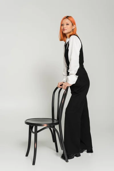 Expressivo e ruivo asiático mulher de cabelos em pé perto da cadeira e olhando para a câmera no fundo cinza, elegante preto e branco roupas, negócio casual, estilo de vida moderno, comprimento total — Fotografia de Stock