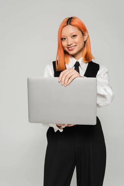 Bonheur, mode de vie freelance, attrayant asiatique femme en noir et blanc vêtements tenant ordinateur portable et souriant à la caméra sur fond gris, cheveux roux colorés, occasionnel d'affaires, génération z — Photo de stock