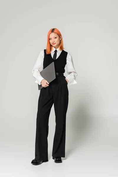 Positive and confident freelancer, young asian woman with red colored hair standing with hand in pocket and laptop on grey background, business casual, white shirt, black vest and pants — Stock Photo