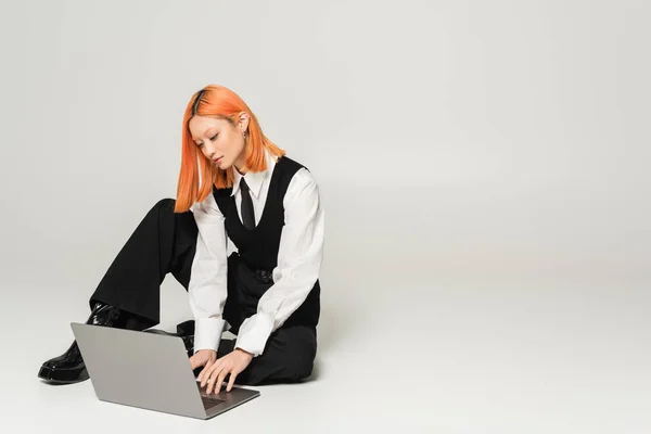 Freelance lifestyle, young asian woman with red dyed hair sitting and working on laptop on grey background, white shirt, black vest, tie, pants, business casual fashion, generation z, full length — Stock Photo