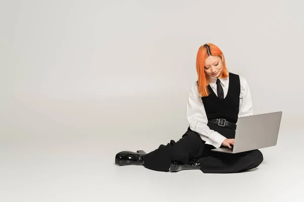 Emoción positiva, cara sonriente, mujer asiática joven en ropa de moda en blanco y negro sentado y trabajando en el ordenador portátil sobre fondo gris, casual de negocios, camisa blanca, pantalones negros y chaleco, freelancer - foto de stock