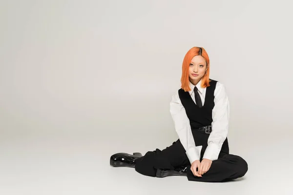 Full length of confident and successful asian woman sitting and looking at camera on grey background, business casual, black vest, tie and pants, white shirt, colored red hair, youthful style — Stock Photo