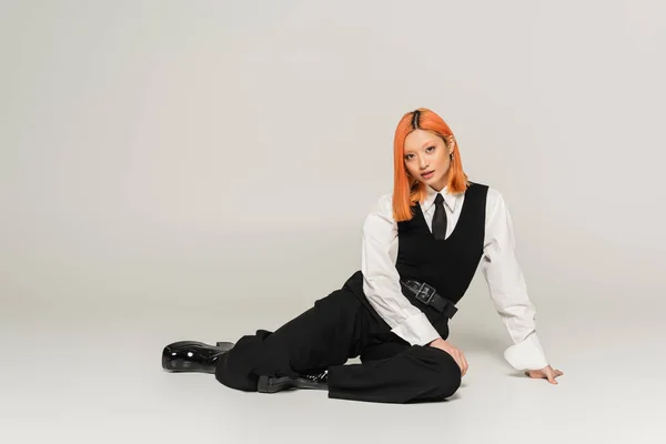Full length of stylish and confident asian woman with dyed red hair sitting and looking at camera on grey background, business casual, black vest and pants, white shirt, modern fashion, gen z — Stock Photo