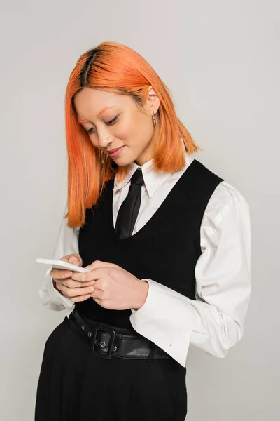 Emoción positiva, cara sonriente, mujer asiática joven en ropa blanca y negra navegar por Internet en el teléfono inteligente sobre fondo gris, pelo rojo de color, camisa blanca, chaleco negro y corbata, casual de negocios - foto de stock