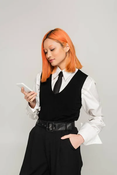 Jeune et élégante femme asiatique avec les cheveux rouges teints debout avec la main dans la poche et en regardant smartphone sur fond gris, chemise blanche, gilet noir, cravate et pantalon, style décontracté d'affaires, gen z — Photo de stock