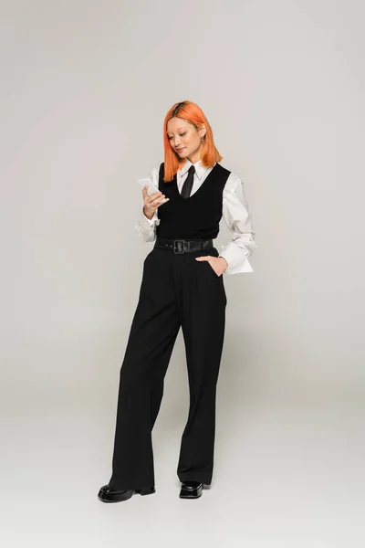 Full length of trendy asian woman with dyed red hair standing with hand in pocket and networking on mobile phone on grey background, business casual fashion, white shirt, black vest and pants — Stock Photo