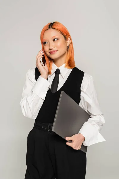 Счастливые эмоции, телефонный звонок, молодая азиатка держит ноутбук и улыбается во время мобильной связи на сером фоне, цветные рыжие волосы, белая рубашка, черный жилет и галстук, деловая повседневная мода — стоковое фото