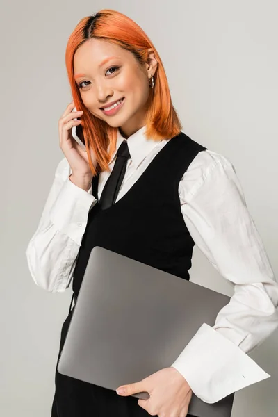 Émotion positive, jeune femme asiatique avec un sourire radieux et les cheveux rouges teints tenant ordinateur portable et parler sur smartphone sur fond gris, chemise blanche, cravate noire et gilet, mode décontractée d'affaires — Photo de stock