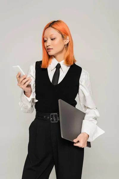 Freelance lifestyle, business casual fashion, red haired asian woman in white shirt, black tie and vest standing with laptop and looking at mobile phone on grey background, generation z — Stock Photo