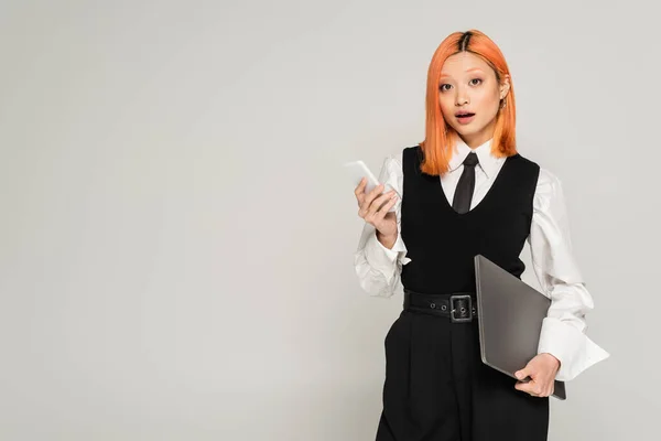 Amazement, surprised asian woman with dyed red hair and open mouth standing with laptop and smartphone while looking at camera on grey background, white shirt, black vest and pants, business casual — Stock Photo