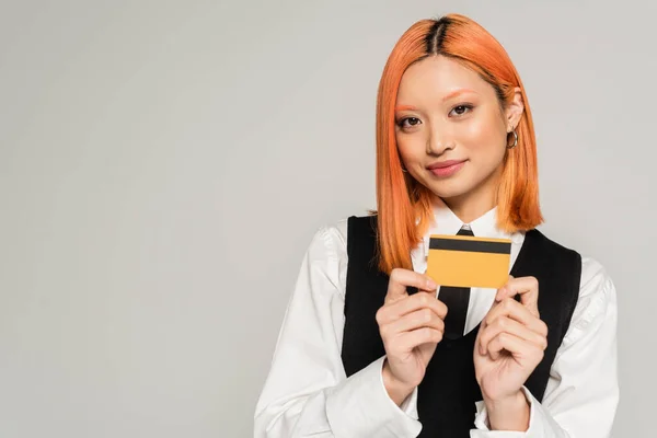 Attractive and positive asian woman with dyed red hair smiling and showing credit card on grey background, white shirt, black vest and tie, business casual fashion, generation z — Stock Photo