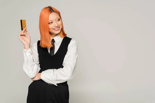 Happy emotion, cheerful smile, red haired asian woman holding credit card and looking away on grey background, business casual style, white shirt, black tie and vest, generation z — Stock Photo