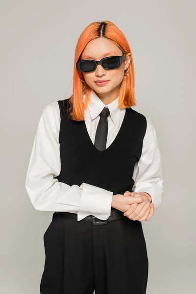 Youthful and fashionable asian woman with colored red hair posing in dark sunglasses, white shirt, black tie and vest on grey background, business casual style, generation z — Stock Photo
