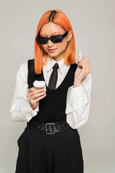 Mujer asiática sonriente con el pelo rojo teñido mirando el caso con auriculares inalámbricos sobre fondo gris, emoción positiva, gafas de sol oscuras, camisa blanca, corbata y chaleco negro, moda casual de negocios - foto de stock