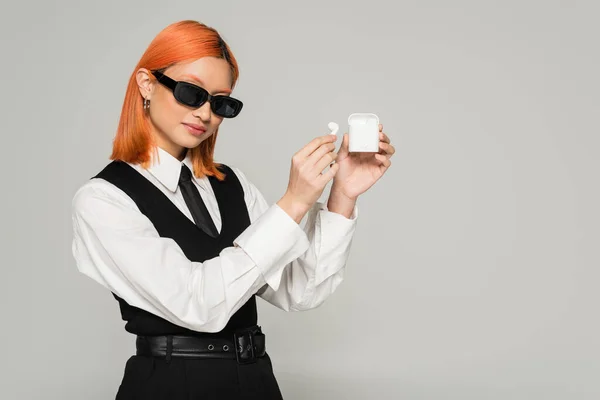 Modern and trendy asian woman in dark sunglasses, with dyed colored hair showing case with earphones on grey background, business casual, white shirt, black tie and vest, generation z lifestyle — Stock Photo