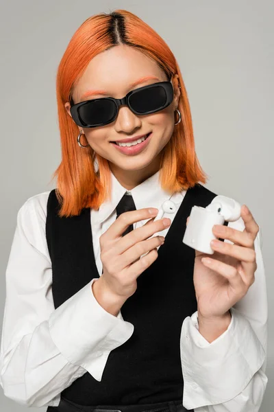 Сверкающая азиатская женщина с сияющей улыбкой и цветными рыжими волосами, держащая футляр с беспроводными наушниками на сером фоне, деловой повседневный стиль, темные солнечные очки, белая рубашка, черный галстук и жилет — стоковое фото