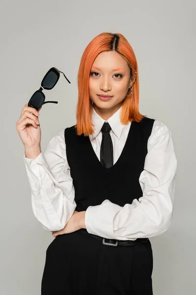 Emoción positiva, sonrisa feliz, mujer asiática joven y de moda con el pelo rojo de color, en camisa blanca, corbata y chaleco negro sosteniendo gafas de sol oscuras y mirando a la cámara en el fondo gris - foto de stock