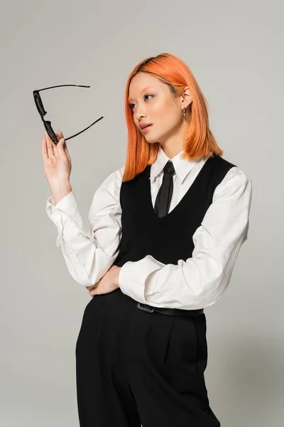 Business casual fashion, attractive asian woman with colored red hair, in white shirt, black pants, vest and tie holding dark sunglasses and looking away on grey background, generation z — Stock Photo