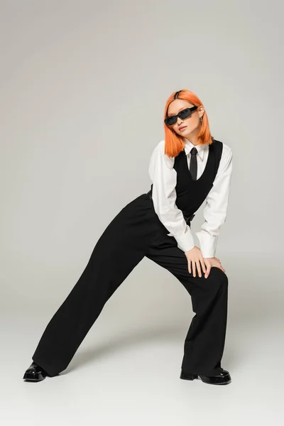 Expressive and young asian woman in dark sunglasses standing in stylish pose on grey background, colored red hair, business casual, black and white clothes, generation z lifestyle, full length — Stock Photo