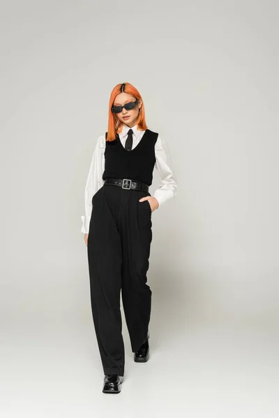 Полная длина современной и модной азиатской женщины в темных солнцезащитных очках и черно-белой одежде ходьба с рукой в кармане на сером фоне, окрашенные рыжие волосы, деловая повседневная мода, современный стиль — стоковое фото
