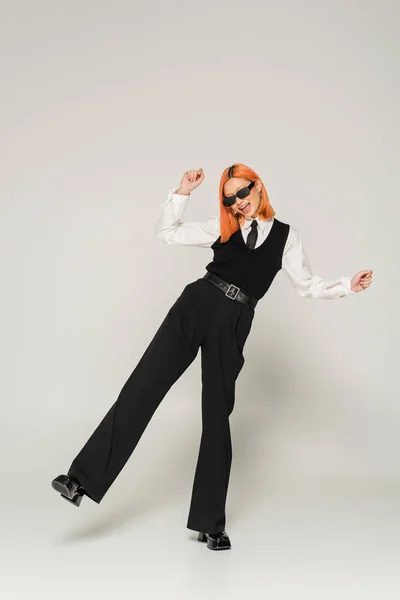 Fröhliche Emotion, überglückliches asiatisches Model posiert mit dunkler Sonnenbrille und trendiger Business-Freizeitkleidung auf grauem Hintergrund, gefärbtem rotem Haar, weißem Hemd, schwarzer Krawatte, Weste und Hose, durchgehende Länge — Stockfoto