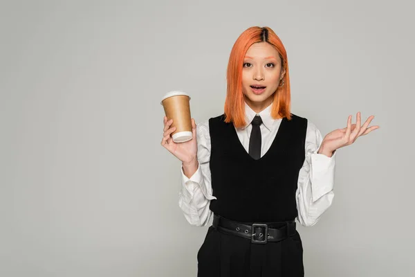 Amazement, surprised asian woman with open mouth holding paper cup, gesturing and looking at camera on grey background, business casual style, white shirt, black tie and vest, colored red hair — Stock Photo