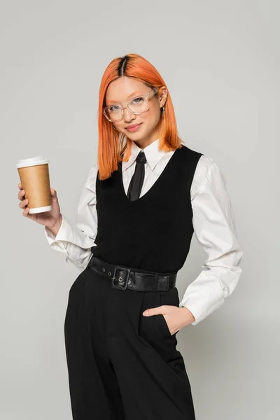 Happy emotion, paper cup with hot takeout drink, red haired asian woman holding hand in pocket and looking at camera on grey background, business casual, stylish eyeglasses, black and white clothes — Stock Photo