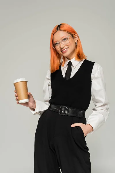 Carefree and red haired asian woman in eyeglasses, white shirt, black tie, vest and pants, with hand in pocket and coffee to go smiling at camera on grey background, generation z lifestyle — Stock Photo