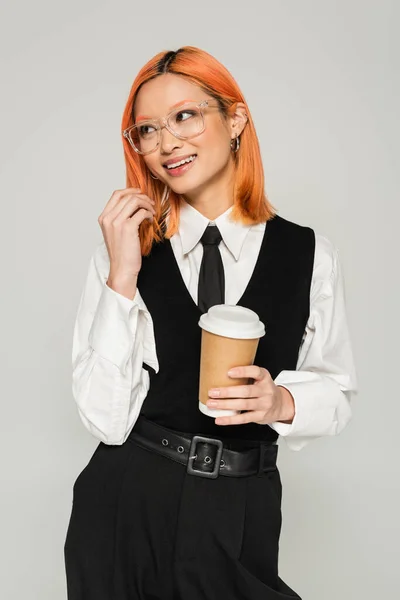 Émotion positive, tasse en papier avec boisson chaude, femme asiatique aux cheveux rouges souriant et regardant loin sur fond gris, cravate noire et gilet, chemise blanche, lunettes élégantes, décontracté d'affaires, génération z — Photo de stock