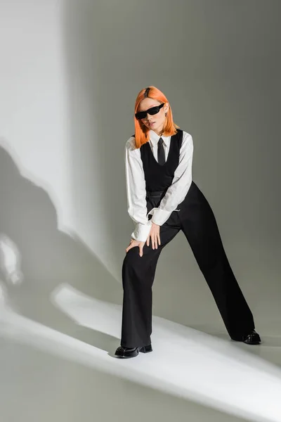Stilvolle Pose einer modischen Asiatin mit gefärbten roten Haaren, dunkler Sonnenbrille, weißem Hemd, schwarzer Krawatte, Weste und Hose auf grauem Hintergrund, Business-Modefotografie, Generation z — Stockfoto