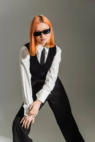 Чорно-білий одяг, темні сонцезахисні окуляри, азіатська жінка з кольоровим червоним волоссям, що стоїть у стильній позі на сірому затіненому фоні, бізнес-мода, покоління Z-життя — Stock Photo