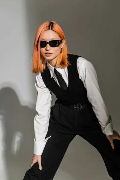 Mujer asiática joven y expresiva con el pelo rojo teñido posando en gafas de sol oscuras, ropa casual de negocios en blanco y negro y mirando a la cámara sobre fondo gris sombreado, generación z - foto de stock