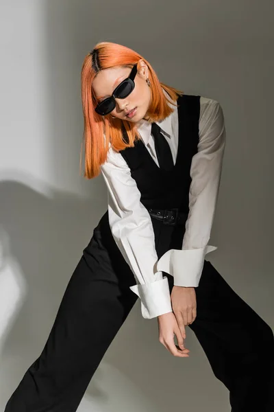 Молодая азиатская модель в темных солнцезащитных очках, стоящая в стильной позе на сером затененном фоне, цветные рыжие волосы, белая рубашка, черный галстук, жилет и брюки, фотография бизнес-моды — стоковое фото