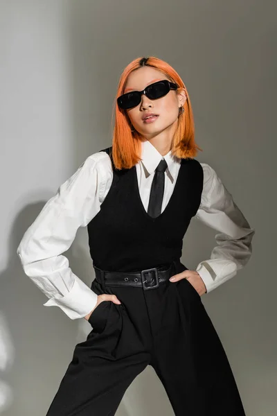Attractive and sensual asian woman with colored red hair posing in dark sunglasses, white shirt, black tie, vest and pants while holding hands in pockets on grey shaded background — Stock Photo