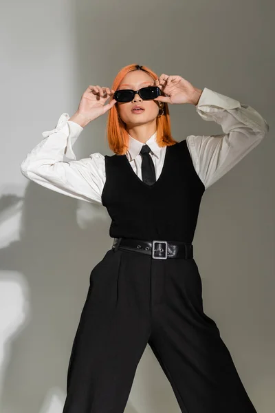 Fashion shoot of expressive asian woman with dyed red hair adjusting dark sunglasses while standing in white shirt, black tie, vest and pants on grey shaded background, business casual style — Stock Photo