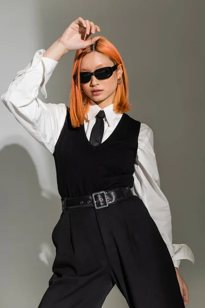 Привлекательная азиатская модель, позирующая с рукой над головой на сером затененном фоне, красные волосы, темные очки, черно-белая деловая повседневная одежда, модная съемка, поколение z — стоковое фото