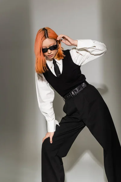 Fashion shoot of modern asian woman in dark sunglasses and black and white clothes on grey shaded background, colored red hair, white shirt, black tie, vest and pants, stylish pose, business casual — Stock Photo