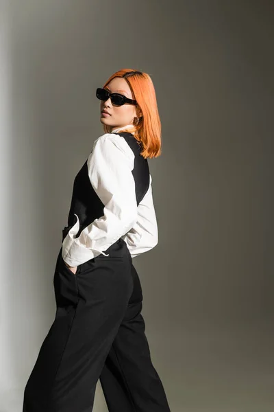 Expressive and red haired asian woman in dark sunglasses, white shirt, vest and pants holding hands in pockets and looking at camera while posing on grey shaded background, modern business fashion — Stock Photo