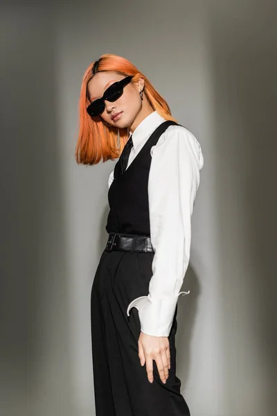 Red haired and expressive asian fashion model looking at camera and posing in dark sunglasses, white shirt and black vest on grey shaded background, business casual clothes, generation z — Stock Photo