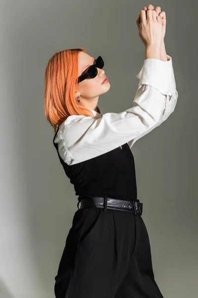 Seitenansicht einer ausdrucksstarken und rothaarigen asiatischen Frau mit dunkler Sonnenbrille, schwarz-weißer Kleidung, die mit erhobenen Händen auf grauem Hintergrund posiert, Business Casual Fashion, Generation Z — Stockfoto
