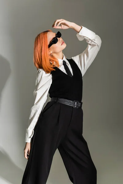 Red haired and expressive asian woman with colored hair posing in dark stylish sunglasses, white shirt, black tie, vest and pants on grey shaded background, business casual fashion, generation z — Stock Photo