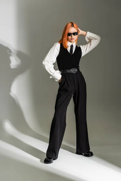 Moda de negocios, mujer asiática de moda con el pelo rojo de color posando con la mano en el bolsillo sobre fondo sombreado gris, gafas de sol oscuras, camisa blanca, corbata negra, chaleco y pantalones, generación z, longitud completa - foto de stock