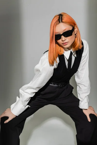 Modern fashion shoot, trendy and red haired asian woman in dark sunglasses and black and white business casual clothes standing in stylish pose on grey shaded background, generation z — Stock Photo