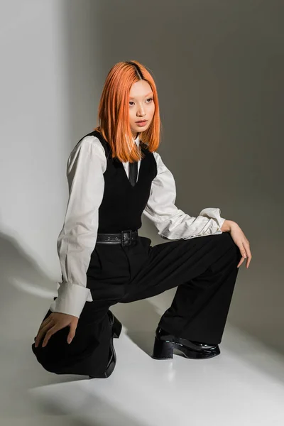 Femme asiatique aux cheveux roux et confiant dans des vêtements de travail élégants posant et regardant loin sur fond gris ombragé, chemise blanche, gilet noir et pantalon, mode moderne, génération z — Photo de stock