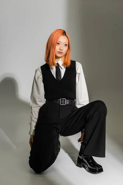 Full length of youthful and stylish asian woman in a white shirt, black vest and pants, with colored red hair posing and looking away on grey shaded background, fashion shoot, generation z — Stock Photo
