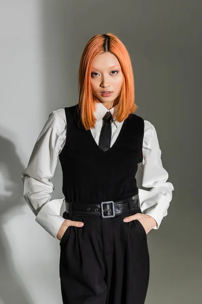 Trendy and confident asian woman with colored hair holding hands in pockets and looking at camera on grey shaded background, white shirt, black tie, vest and pants, business casual fashion — Stock Photo