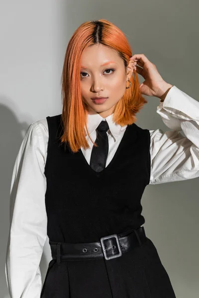 Mujer asiática joven y segura de sí misma fijando el pelo rojo mientras mira a la cámara en el fondo sombreado gris, estilo casual de negocios, ropa en blanco y negro, estilo de vida de generación z, sesión de fotos de moda - foto de stock