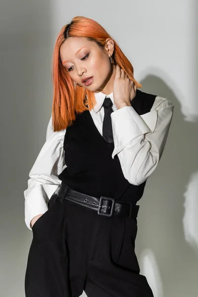 Casual business fashion, attractive asian woman with colored red hair touching neck and holding hand in pocket on grey shaded background, white shirt, black tie, vest and pants, modern lifestyle — Stock Photo