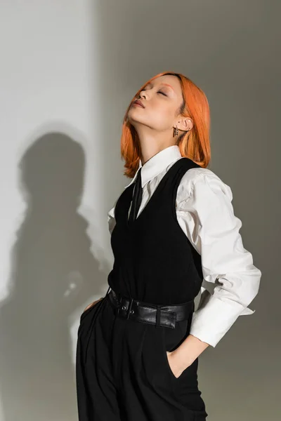 Чувственная и модная азиатская женщина с красными рыжими волосами, закрытыми глазами и руками в карманах, позирующими в белой рубашке, черном жилете, брюках и галстуке на сером затененном фоне, повседневная деловая мода — стоковое фото