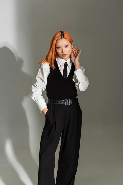 Mujer asiática de moda y confiada tocando el pelo de color rojo, sosteniendo la mano en el bolsillo y mirando a la cámara sobre fondo gris sombreado, casual de negocios, camisa blanca, corbata negra, camisa y pantalones - foto de stock